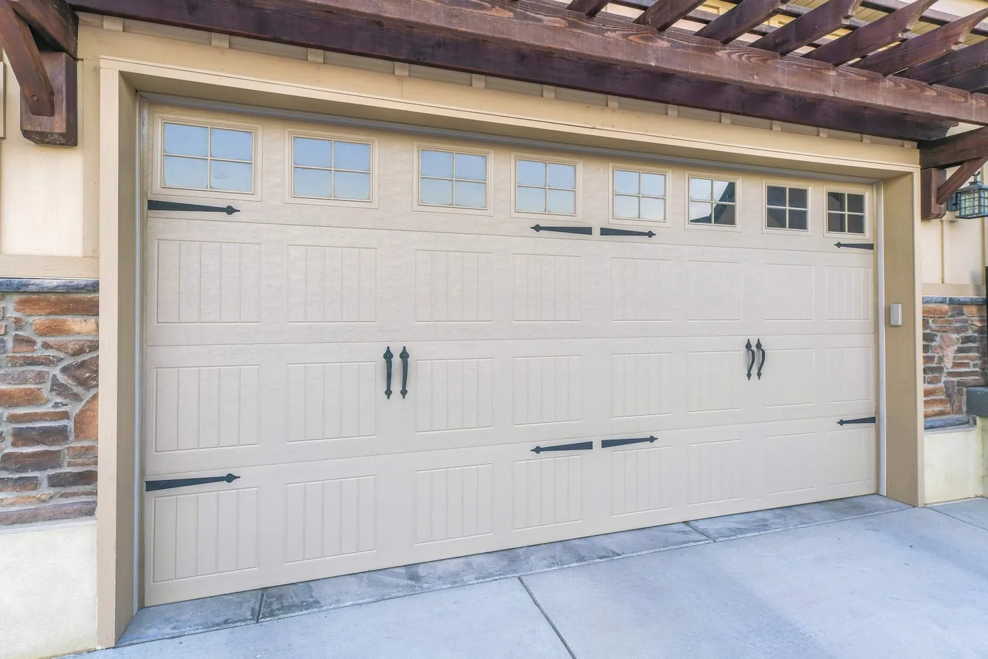 Cream carriage-style garage door with upper windows and black decorative hardware beneath a wood pergola.