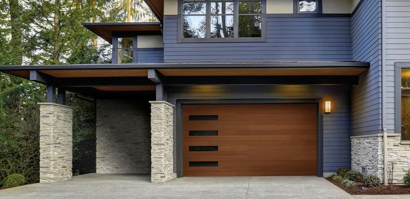 Modern wood-look garage door with offset windows on a gray home with stone columns.